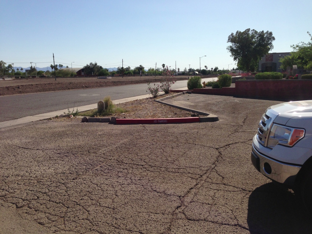 We Do Lines Arizona - A cracked asphalt parking lot with a single red parking barrier marked "visitor" near the center features faded line striping. A partial view of a white vehicle is visible on the right. Sparse vegetation and distant buildings are in the background.
