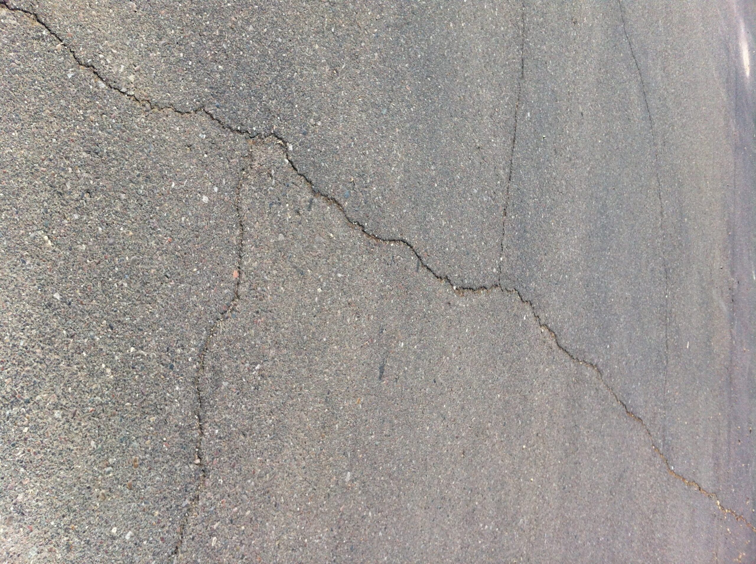 We Do Lines Arizona - A close-up of an asphalt surface with several noticeable cracks running across it, interrupted by faded line striping.