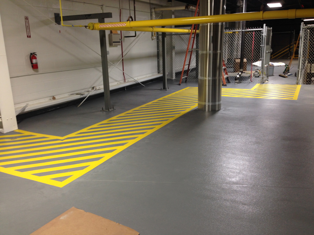 We Do Lines Arizona - A factory or warehouse floor with a shiny grey surface, featuring precise line striping in yellow and black safety markings, surrounded by structural pipes and metal fencing.