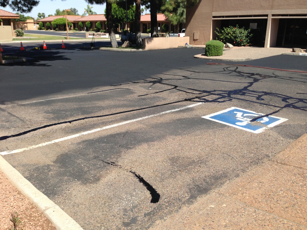 We Do Lines Arizona - A parking lot with visible cracks in the asphalt. An accessible parking space is marked in fresh line striping on the right side. Three orange traffic cones are positioned in the background.