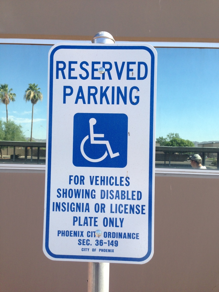 We Do Lines Arizona - Sign indicating "Reserved Parking" for vehicles with a disabled insignia or license plate, as per Phoenix City Ordinance Sec. 36-149. The line striping is clearly visible, with palm trees and buildings reflected in the background.