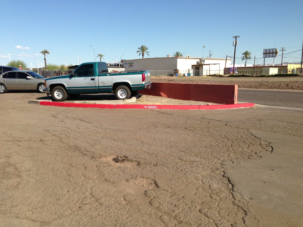 We Do Lines Arizona - A green and white pickup truck is parked atop a red curb in a nearly empty parking lot on a sunny day. Line striping clearly marks the spaces, with palm trees and industrial buildings visible in the background.