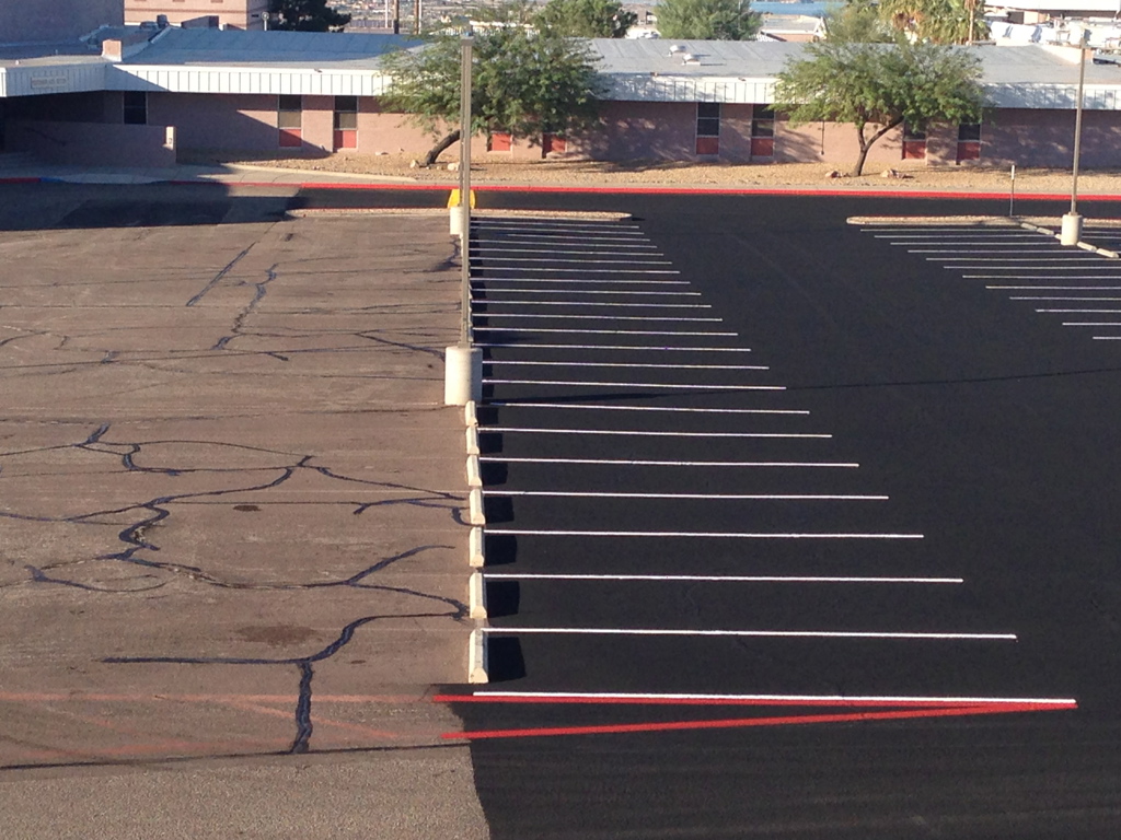 We Do Lines Arizona - A parking lot is shown with newly resurfaced asphalt on the right side and older cracked pavement on the left, marked with white line striping. In the background are trees and buildings.