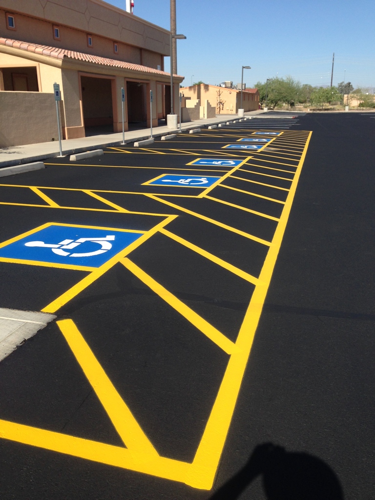 We Do Lines Arizona - A row of freshly painted disabled parking spaces in a parking lot outside a building under a clear sky showcases precise line striping. Yellow lines and blue wheelchair symbols are clearly visible on the asphalt.