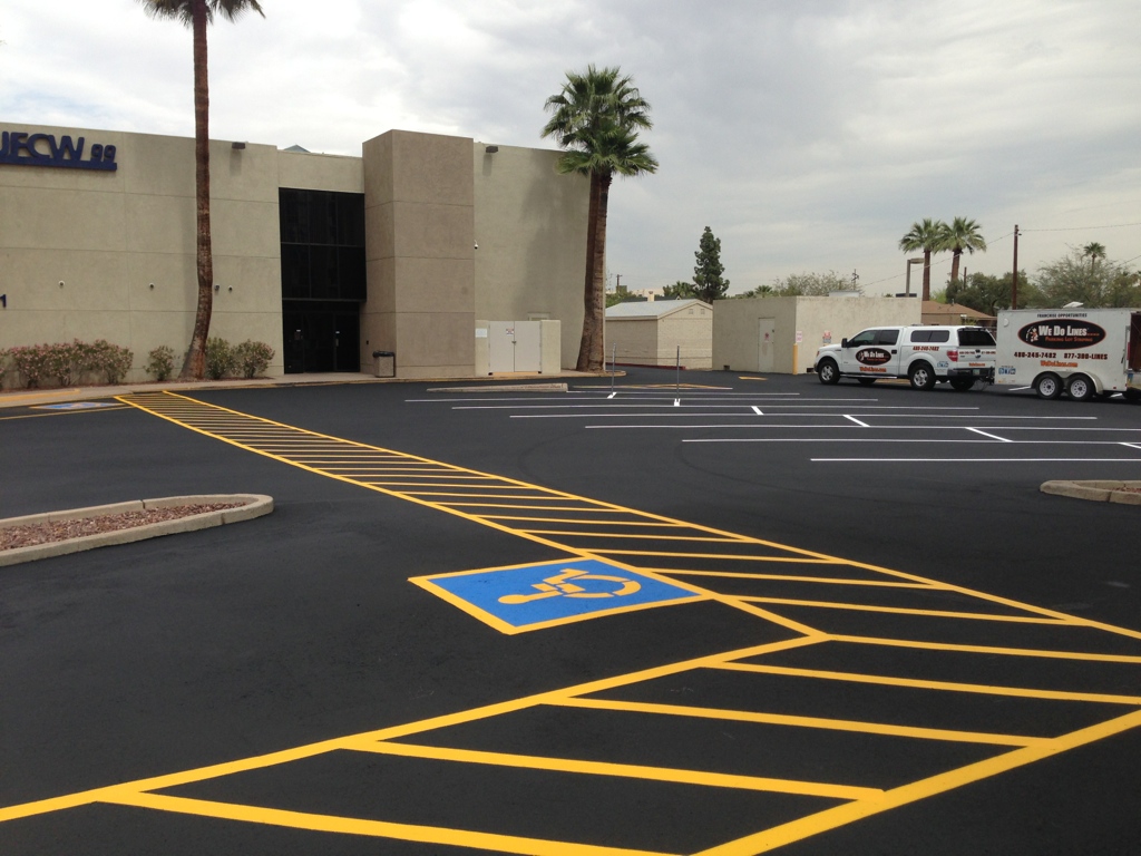 We Do Lines Arizona - A parking lot with freshly painted line striping, including a disabled parking space marked in blue and yellow. Two parked vehicles and a trailer are seen near a beige building with palm trees around.