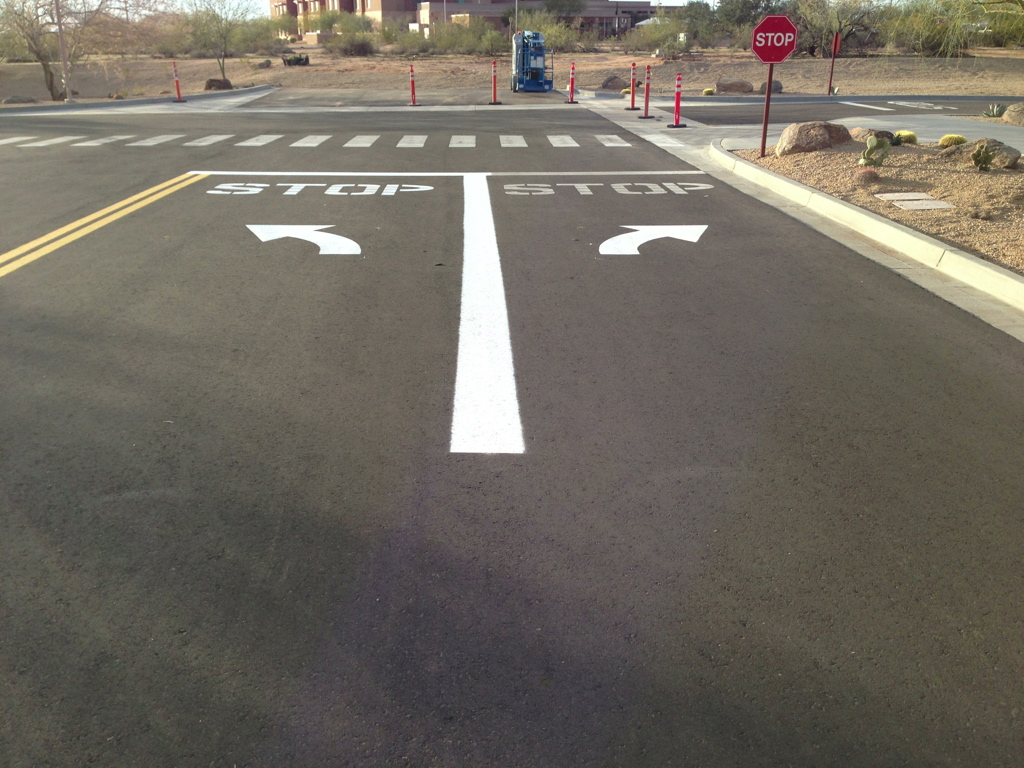 We Do Lines Arizona - A newly paved road with crisp line striping, featuring clearly marked white directional arrows and "STOP" signs painted on the asphalt, leads to a crosswalk and a stop sign at the intersection.