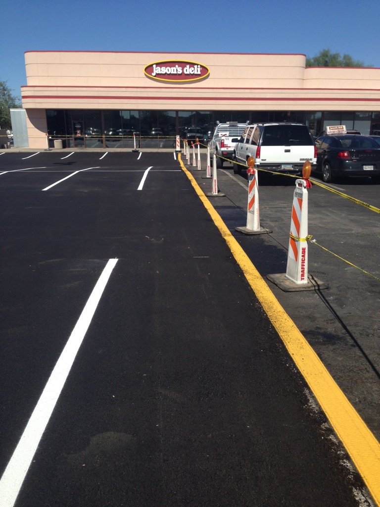 We Do Lines Arizona - A recently paved parking lot with fresh white lines and yellow curbs in front of a Jason's Deli showcases meticulous line striping. Several traffic cones and caution tape are placed along the edge.