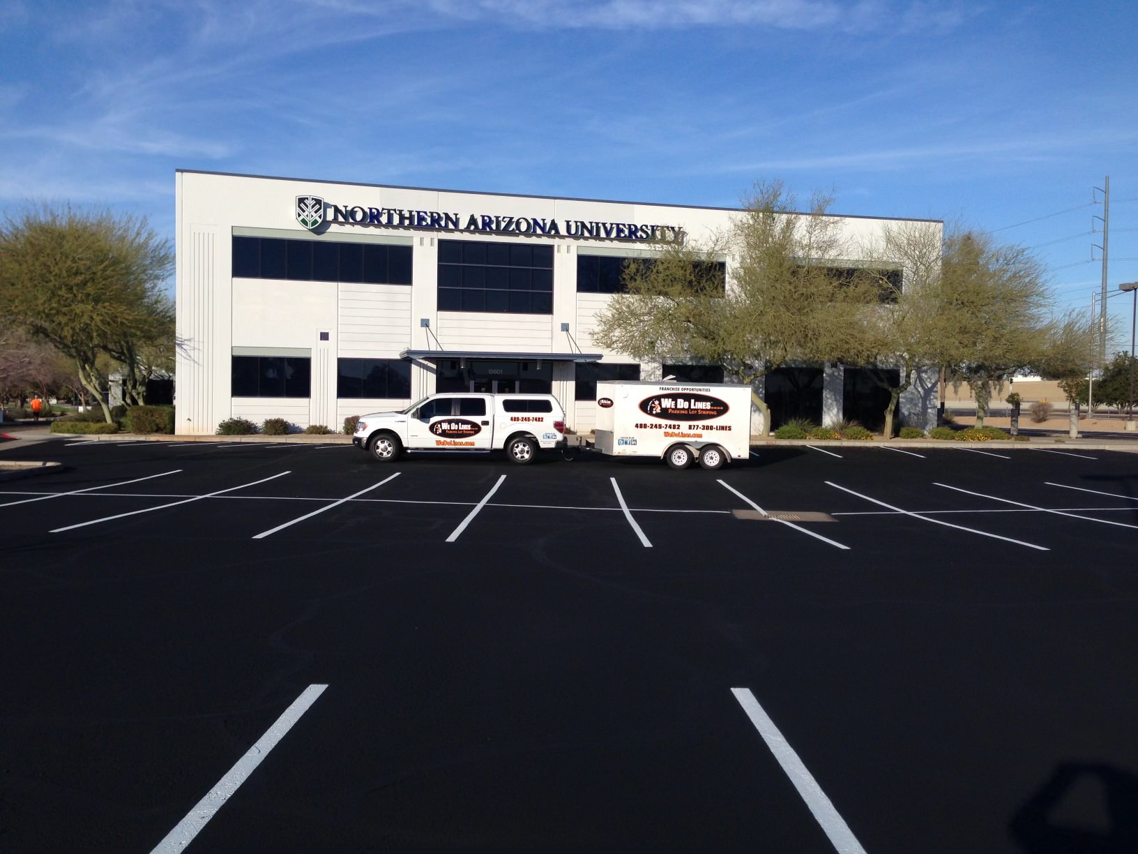 We Do Lines Arizona - Two vehicles and a trailer parked in front of a building labeled Northern Arizona University with freshly done line striping in the empty parking lot and clear skies above.