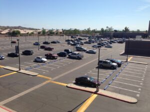 We Do Lines Arizona - An expansive parking lot with numerous parked cars is seen on a sunny day, its line striping neatly defining each space. A building is visible in the distance, with trees and houses appearing beyond the parking area.