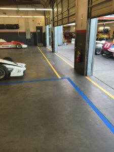 We Do Lines Arizona - A garage interior with open bay doors and racing cars parked inside, including a white and red race car labeled "17" in one of the bays. The floor features precise line striping with marked blue and yellow lines.