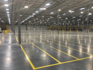 We Do Lines Arizona - A large, empty warehouse with a high ceiling, concrete floor, and meticulously line striping in yellow on the ground. Metal shelves line the far wall, and the space is well-lit with ceiling lights.