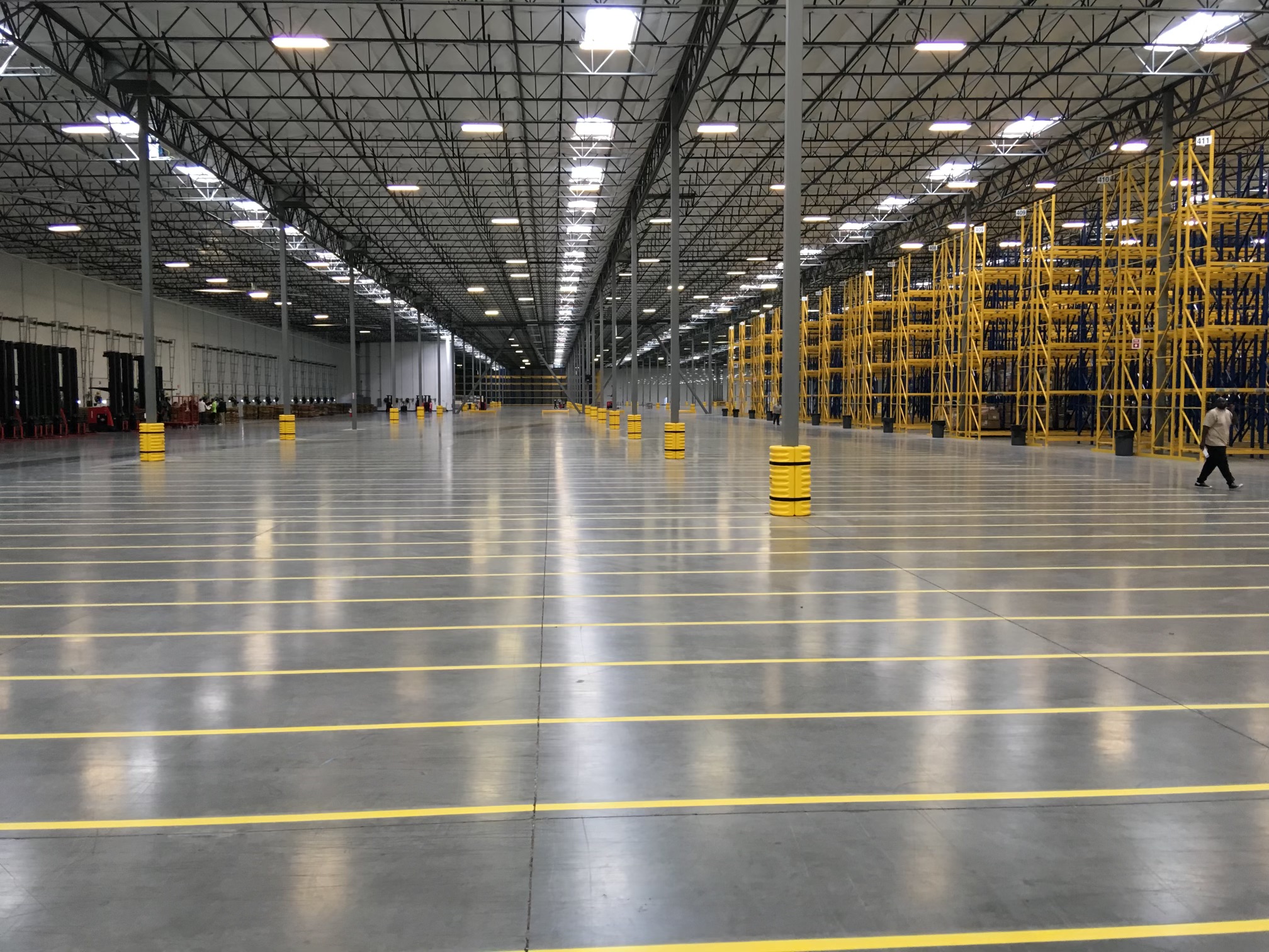 We Do Lines Arizona - A spacious warehouse interior with high ceilings, orderly rows of shelving units, yellow line striping on the floor, and bright overhead lighting. Few individuals are visible in the distance.