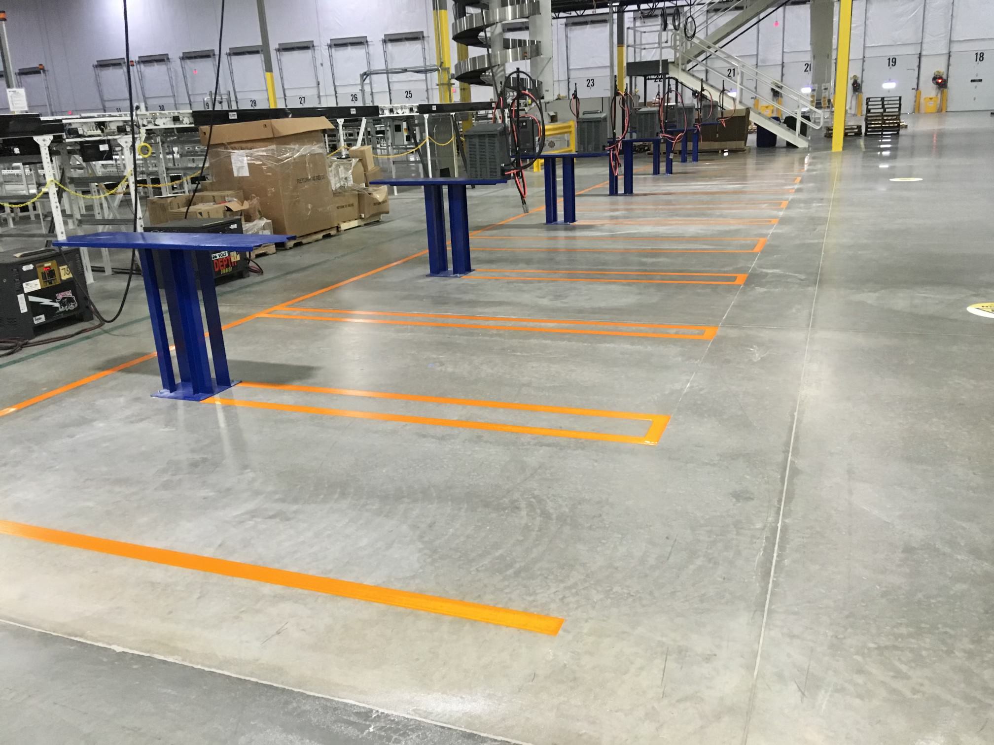 We Do Lines Arizona - Empty workstation area in an industrial facility with blue equipment stands and orange line striping on the floor. In the background are stairs and other machinery.