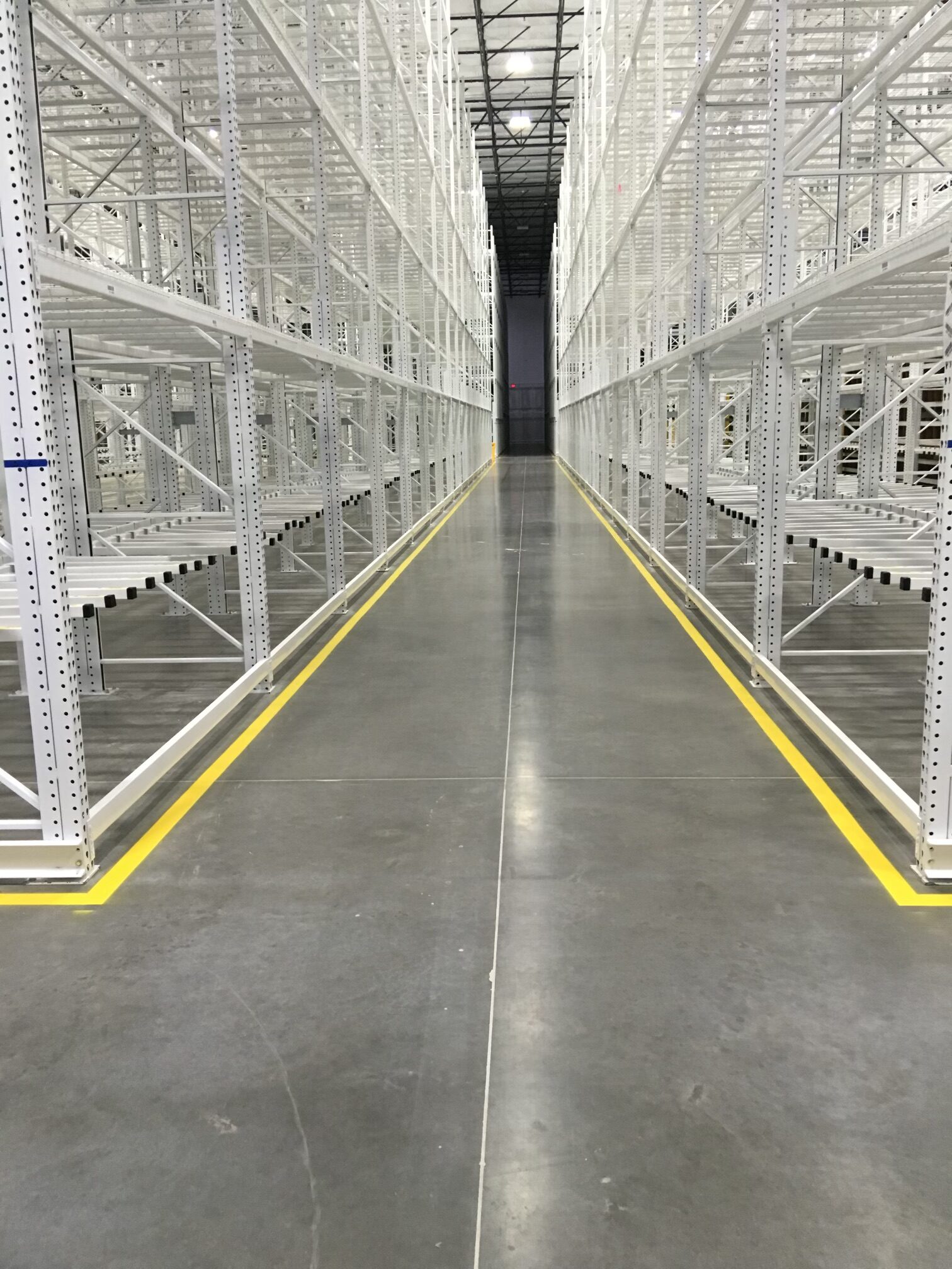 We Do Lines Arizona - A view down an empty aisle in a large, well-lit warehouse with metal shelving units on both sides and yellow line striping marking the pathway on the polished concrete floor.