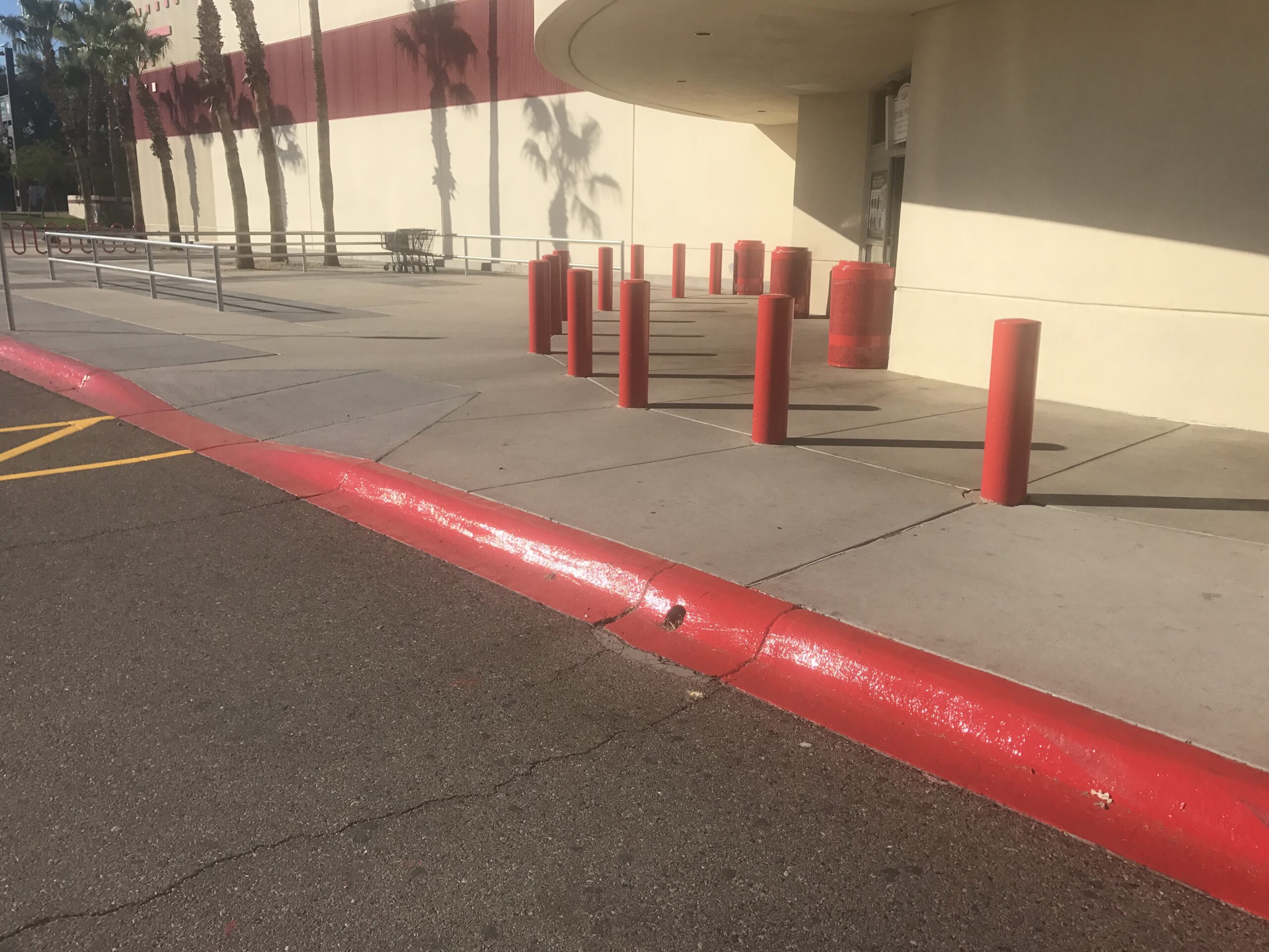 We Do Lines Arizona - A sidewalk with red bollards and a ramp leading to the entrance of a beige building, complemented by meticulous line striping along the red-painted curb. Palm tree shadows are cast on the building.