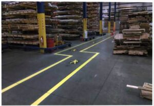 We Do Lines Arizona - A warehouse interior with organized shelving and stacks of materials. Line striping in yellow, along with pedestrian symbols, indicates walking paths through the area.